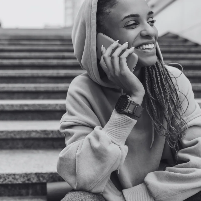 JTL_BW-Options-02 A black-and-white image of a smiling person wearing a hoodie and a smartwatch, sitting on stairs while holding a phone to their ear.