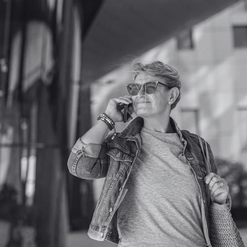 JTL_BW-Options-04 A black-and-white image of a person wearing sunglasses and a denim jacket, talking on the phone while standing outside a modern building, carrying a woven bag.