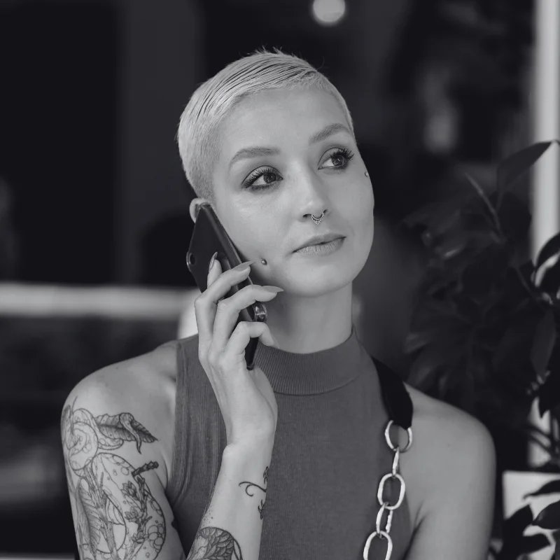 JTL_BW-Options-05 A black-and-white image of a person with short-cropped hair, piercings, and tattoos, holding a phone to their ear, wearing a sleeveless top with chain accents, and looking thoughtfully to the side.
