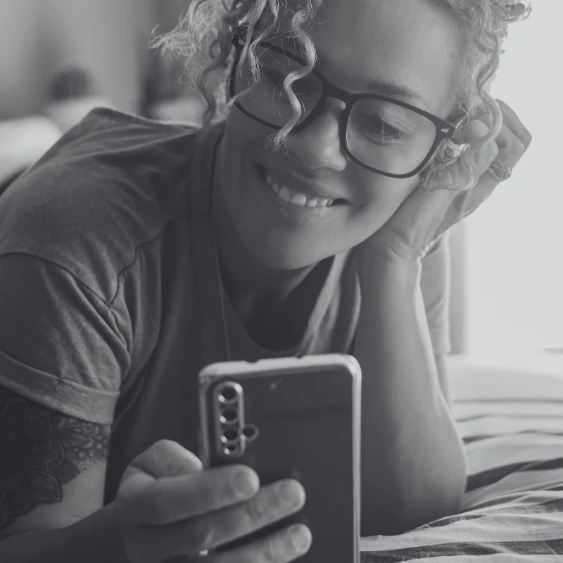 JTL_BW-Options-07 A black-and-white image of a person with curly hair and glasses smiling while looking at their phone. They are lying down with one hand resting on their head.