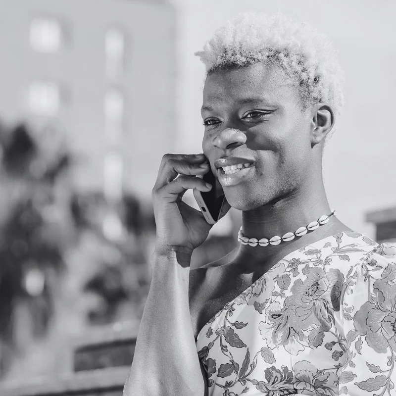JTL_BW-Options-08 A black-and-white image of a smiling person with short, light-colored hair wearing a floral-patterned top and a shell necklace, talking on a cellphone outdoors.