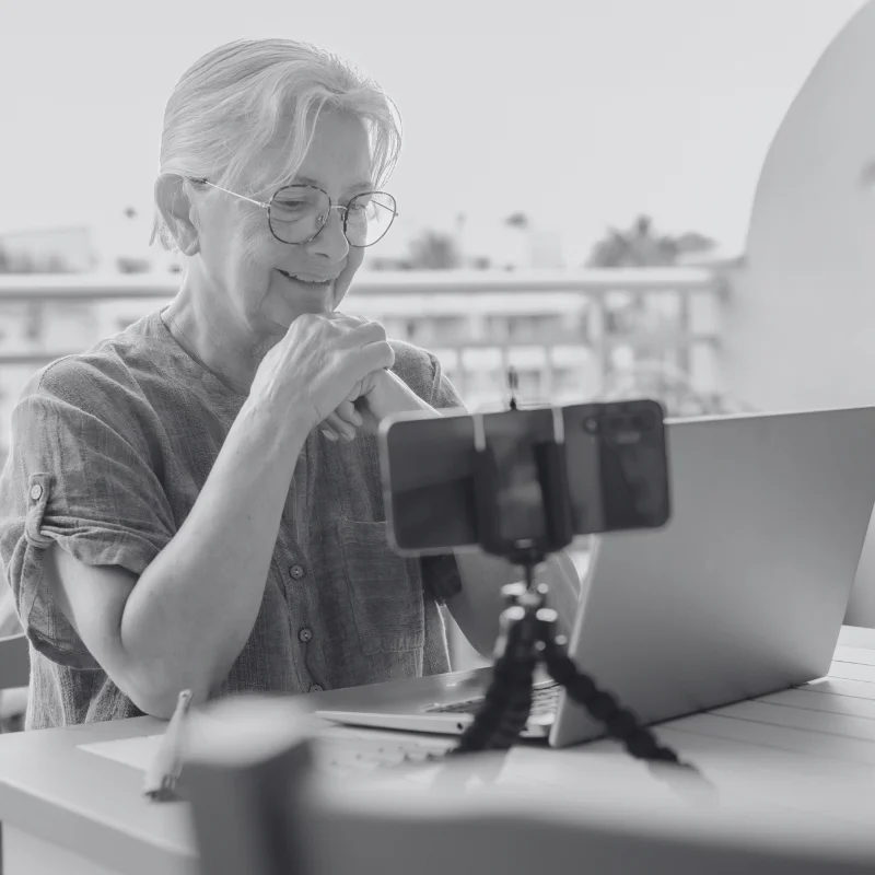 JTL_BW-Options-09 An elderly woman with white hair and glasses, wearing a light blouse, sitting on a balcony, smiling at a smartphone mounted on a tripod, with a laptop on the table.