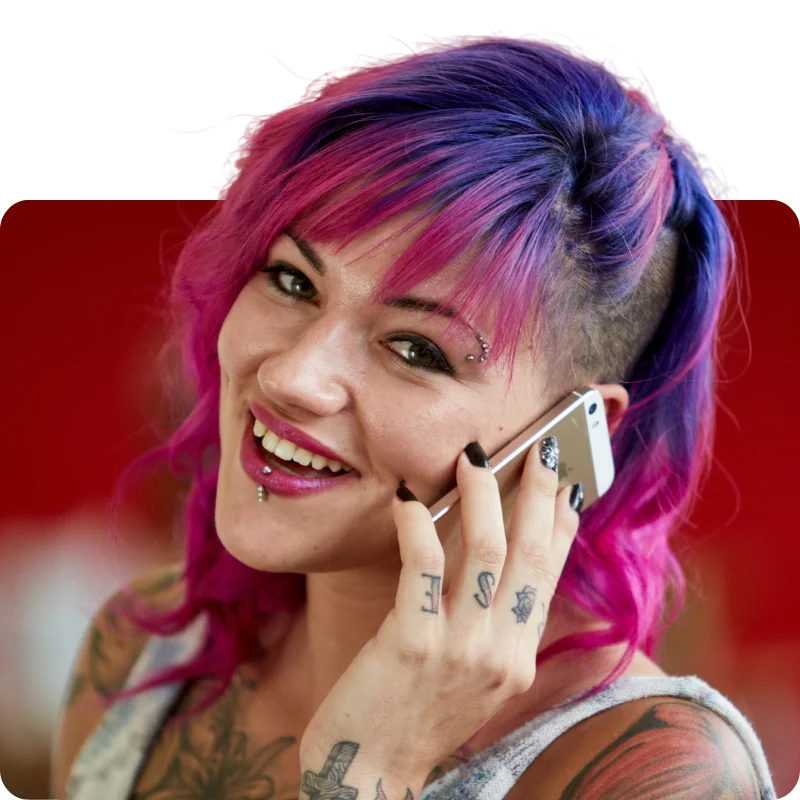 A person with vibrant pink and purple hair styled with a shaved side, wearing a tank top, tattoos visible on their arms and hand, smiling while talking on a smartphone.