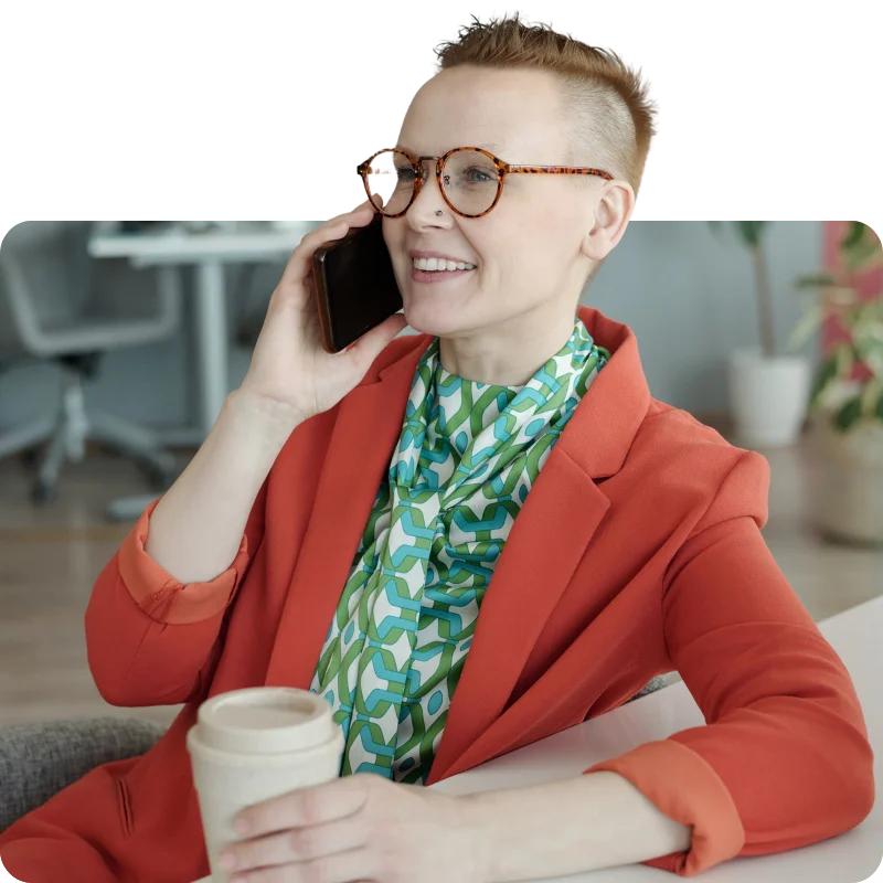 JTL_Cropped-Options-011 A person with short, styled hair wearing an orange blazer and patterned shirt, holding a coffee cup while smiling and talking on a smartphone in a modern office setting.