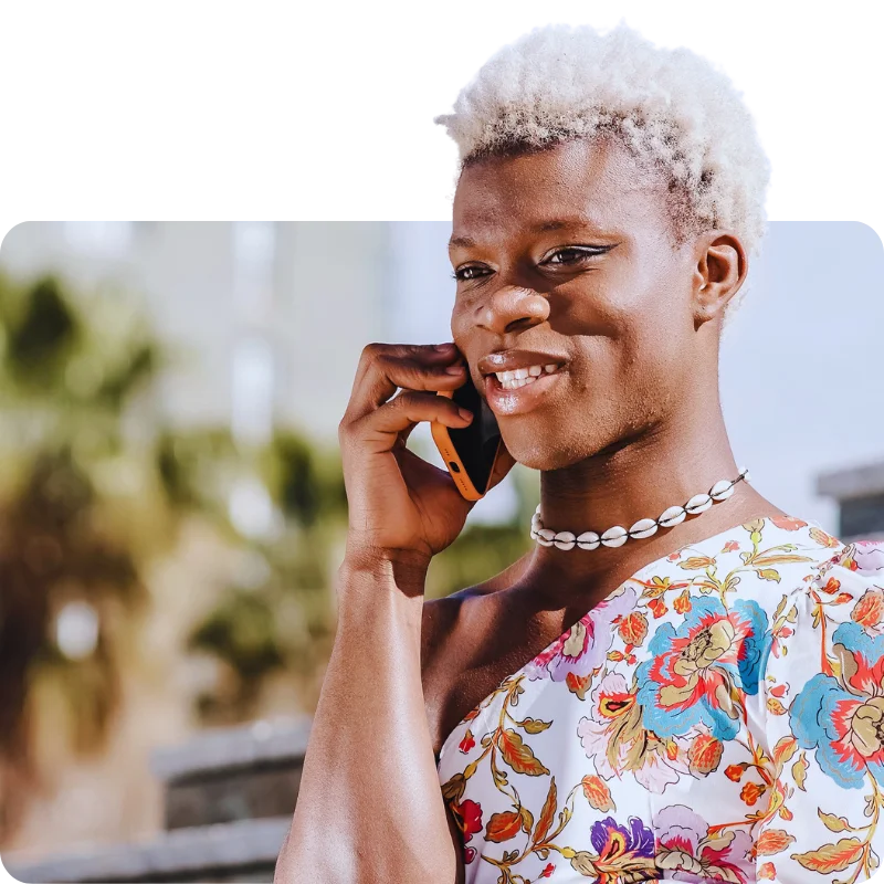 JTL_Cropped-Options-012 A smiling person with short, bleached hair wearing a floral-patterned outfit and a shell necklace, talking on a smartphone outdoors with blurred greenery and urban surroundings in the background.