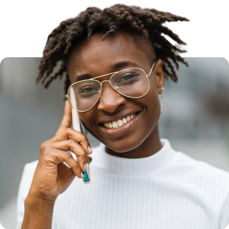 JTL_Cropped-Options-013 A person with a cheerful expression, short textured hair, and stylish glasses, wearing a white shirt, talking on a smartphone outdoors with a blurred urban background.