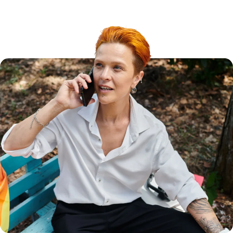 A person with short orange hair, wearing a white shirt and sitting on a colorful bench, talking on a smartphone in an outdoor setting surrounded by trees.