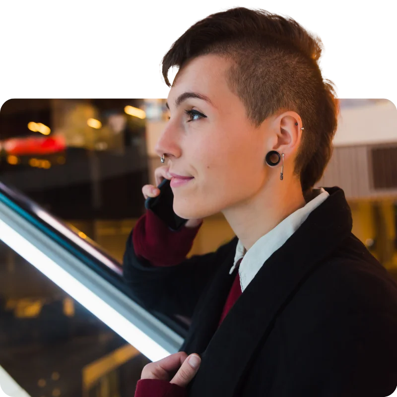 JTL_Cropped-Options-015 A person with a side-shaved hairstyle and earrings, wearing a black coat and red sweater, talking on a smartphone while standing near an escalator in a brightly lit indoor setting.