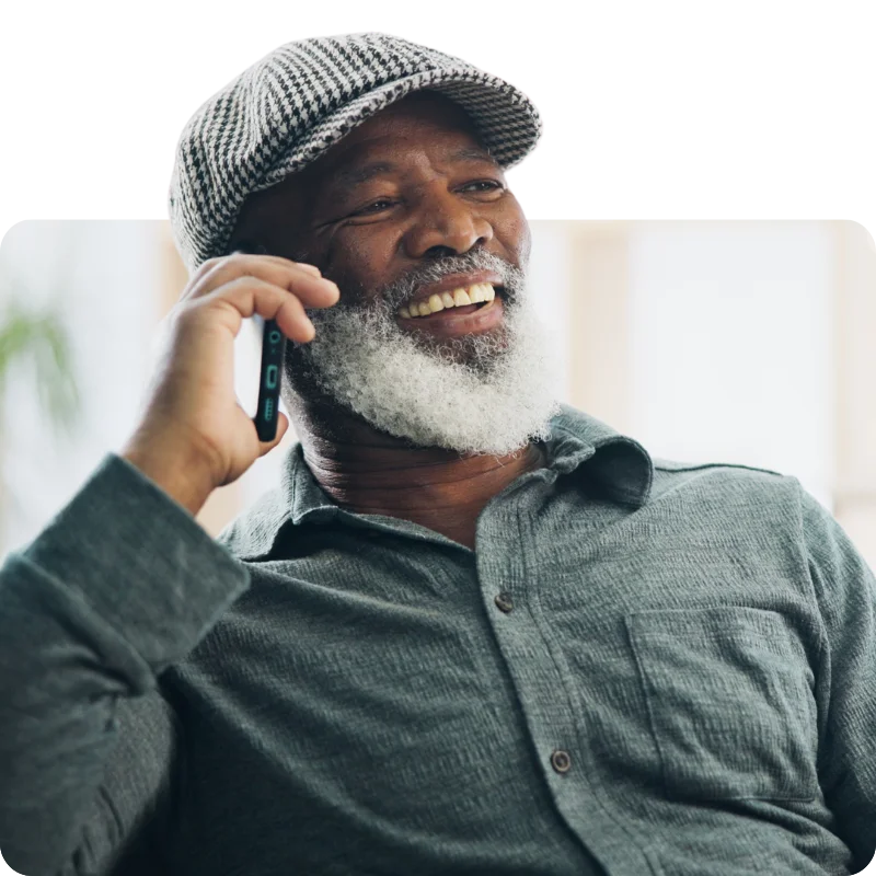 JTL_Cropped-Options-016 A smiling elderly man with a white beard wearing a checkered cap and a gray shirt, engaged in a phone conversation.
