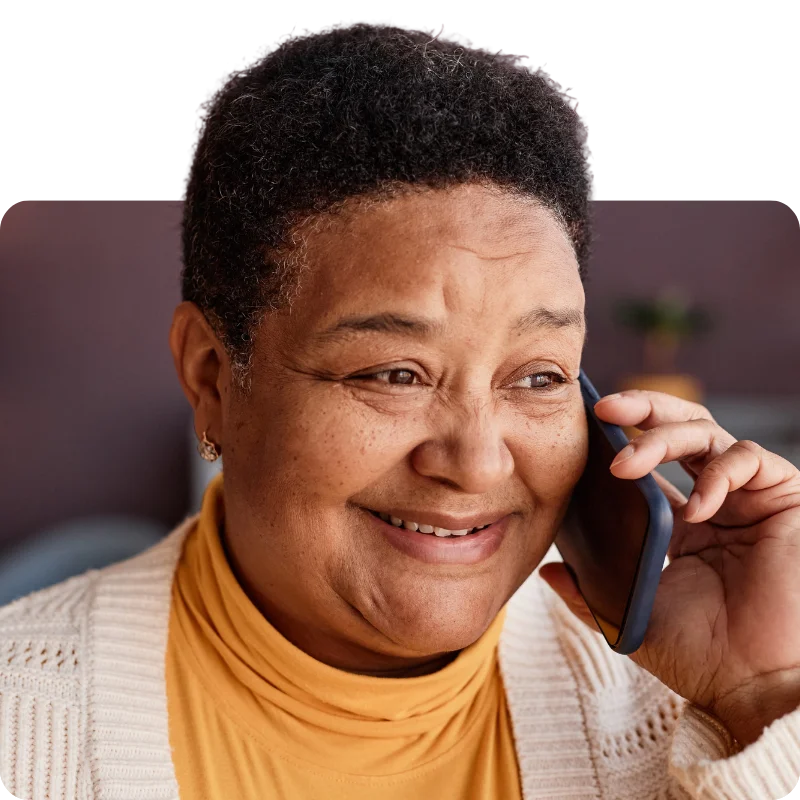 JTL_Cropped-Options-017 A cheerful elderly woman wearing a yellow turtleneck and cream cardigan, smiling warmly during a phone conversation.