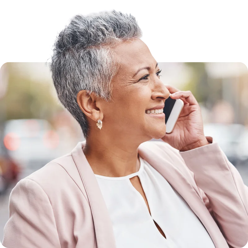 A confident elderly woman with short gray hair, wearing a white top and pink blazer, smiling as she talks on her smartphone outdoors.