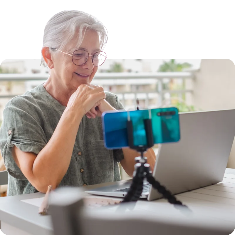 JTL_Cropped-Options-019 An elderly woman with white hair and glasses, wearing a green blouse, sitting on a balcony while using a laptop and smiling at a smartphone mounted on a tripod.