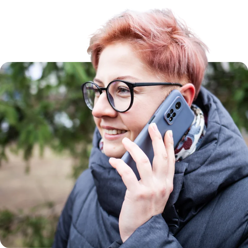 JTL_Cropped-Options-04 A smiling person with short pink hair and glasses, wearing a scarf and coat, speaking on a smartphone outdoors.