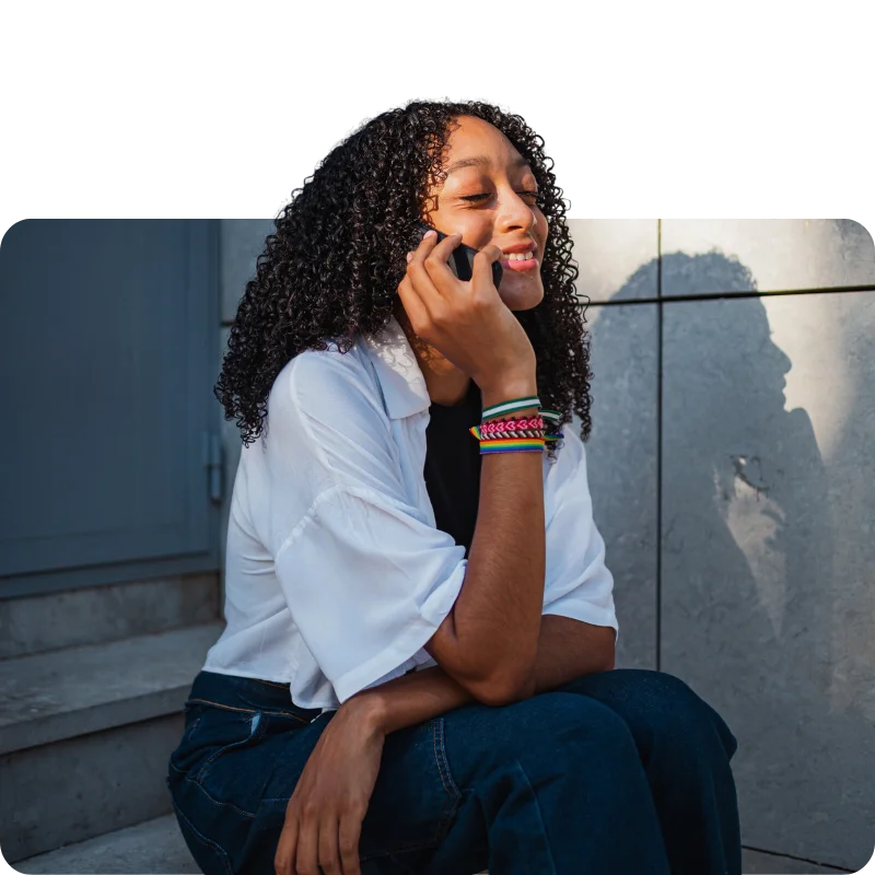 JTL_Cropped-Options-05 A person with curly hair, wearing a white shirt and colorful bracelets, sitting outdoors on steps while talking on the phone with a cheerful expression.