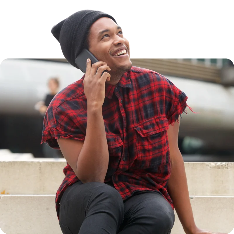 JTL_Cropped-Options-06 A person in a red flannel shirt and black beanie sitting on steps, smiling warmly while talking on the phone outdoors.