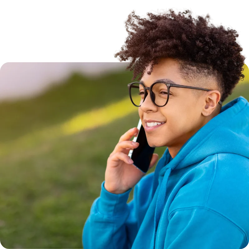 JTL_Cropped-Options-07 A person wearing glasses and a bright blue hoodie, sitting on grass, smiling while talking on the phone.