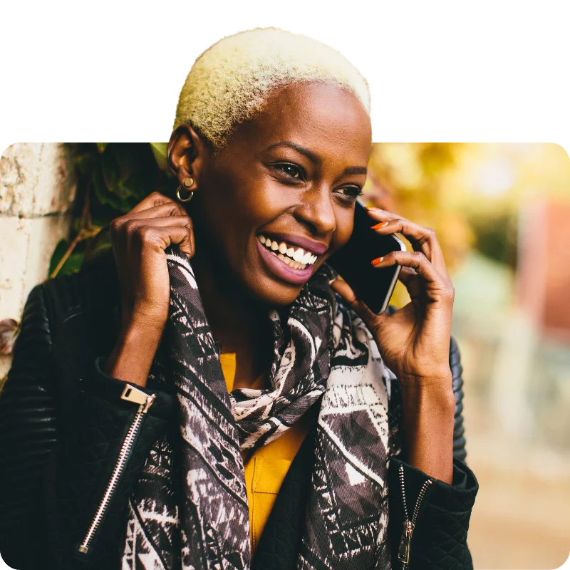 JTL_Cropped-Options-08 A person with short blonde hair, wearing a patterned scarf and a leather jacket, smiling brightly while talking on the phone outdoors.
