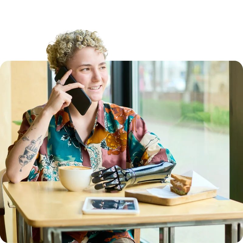JTL_Cropped-Options-09 A person with short curly hair, wearing a colorful floral shirt, sitting at a café table while talking on the phone, with a cup of coffee and a prosthetic arm visible.